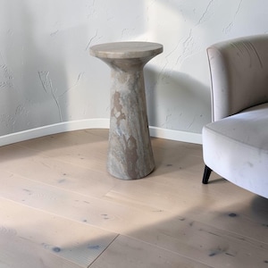 Puede incluir: Una mesa auxiliar cil&iacute;ndrica hecha de piedra natural con una superficie circular lisa y una base c&oacute;nica. La piedra presenta un patr&oacute;n variado de remolinos en tonos beige, tostado y gris en toda su superficie, creando un aspecto marmolado.