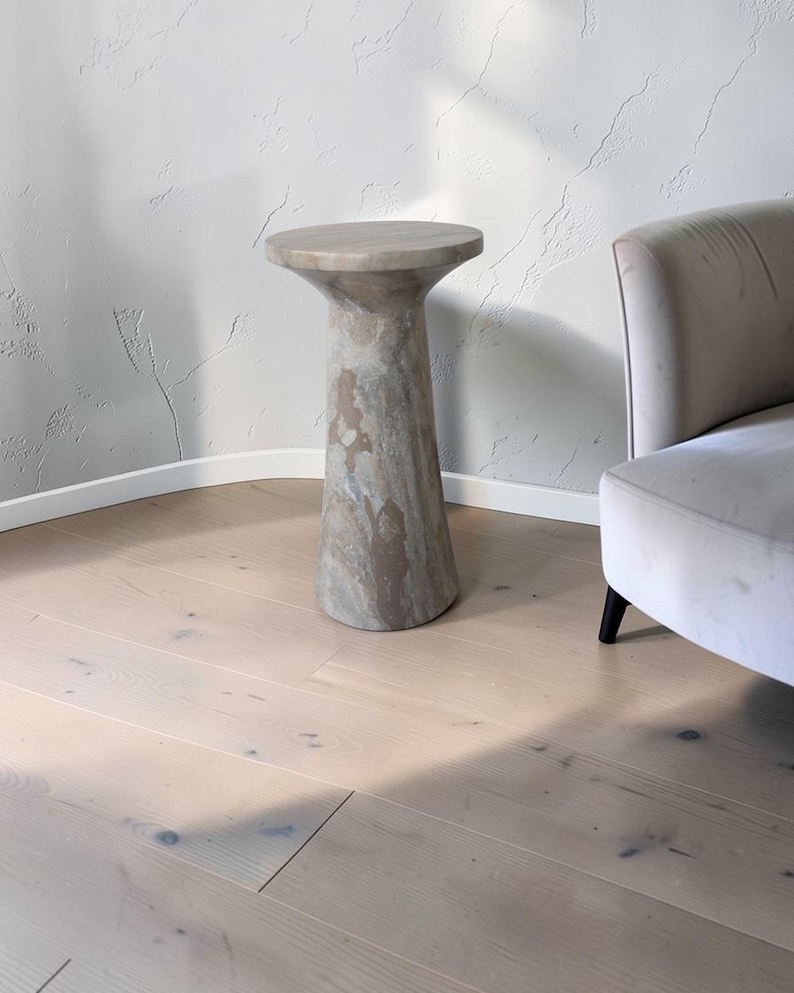 Puede incluir: Una mesa auxiliar cil&iacute;ndrica hecha de piedra natural con una superficie circular lisa y una base c&oacute;nica. La piedra presenta un patr&oacute;n variado de remolinos en tonos beige, tostado y gris en toda su superficie, creando un aspecto marmolado.