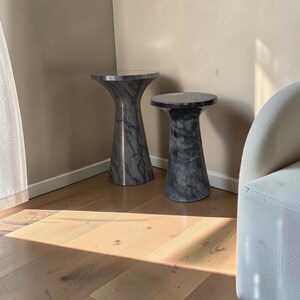 Puede incluir: Dos mesas auxiliares cil&iacute;ndricas con un acabado de m&aacute;rmol gris oscuro y negro. Cada mesa presenta una superficie superior circular lisa que se estrecha hacia una base m&aacute;s ancha y estable, mostrando un patr&oacute;n de piedra uniforme en toda la estructura.