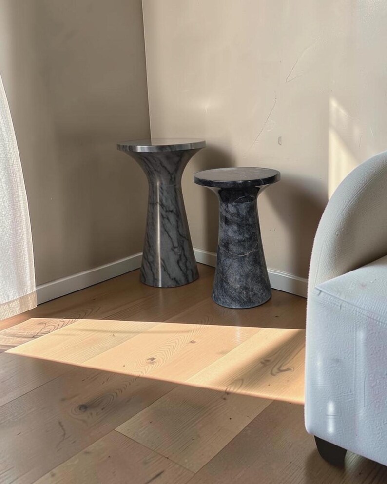Puede incluir: Dos mesas auxiliares cil&iacute;ndricas con un acabado de m&aacute;rmol gris oscuro y negro. Cada mesa presenta una superficie superior circular lisa que se estrecha hacia una base m&aacute;s ancha y estable, mostrando un patr&oacute;n de piedra uniforme en toda la estructura.
