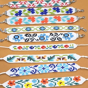May include: A collection of nine woven bracelets featuring various colorful cross-stitch patterns. The designs include floral motifs, geometric shapes, and abstract patterns in shades of red, blue, green, yellow, and black on white backgrounds.