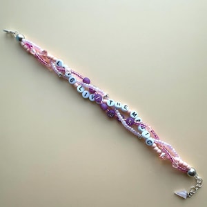 May include: A multi-strand beaded bracelet featuring a mix of light pink, dark pink, and purple beads. The bracelet includes white letter beads spelling out the phrase "LVG THE MAGIC" interspersed with small clear crystals and silver-tone metal accents.