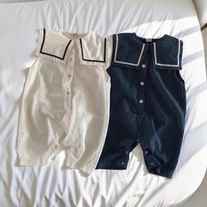May include: Two baby rompers. One is off-white with a black-trimmed sailor collar, and the other is navy blue with a white-trimmed sailor collar. Both have button closures down the front and at the legs. The rompers are sleeveless.