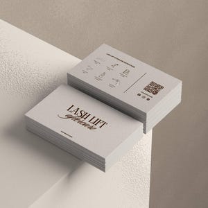 以下が含まれることがあります： 「LASH LIFT aftercare」の文字がエレガントな筆記体で書かれた、明るい灰色の名刺の束。一番上の束には、まつげリフトのアフターケアの説明とQRコードが表示されています。カードは、ニュートラルな背景の白い表面に置かれています。