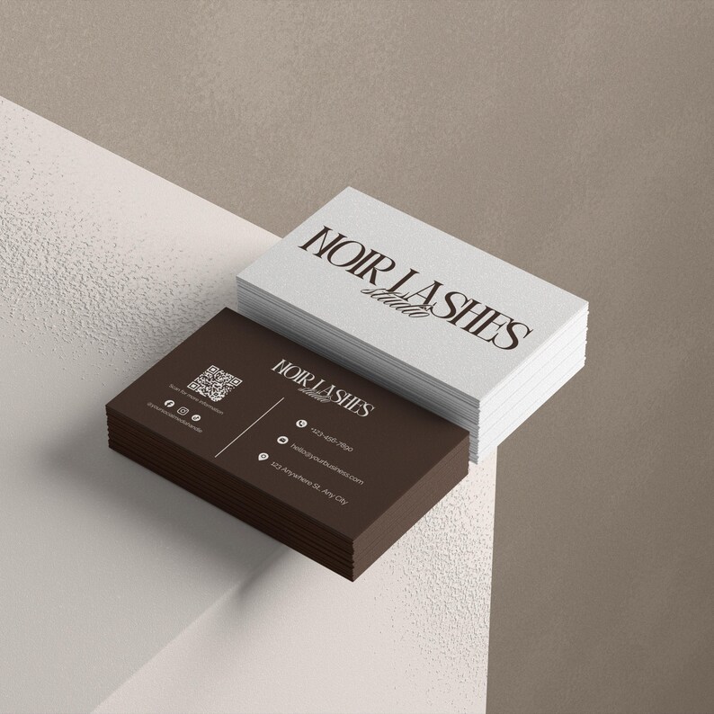 May include: A stack of business cards with the text "NOIR LASHES Studio" in a stylish font. The bottom card is dark brown with contact information and a QR code. The top cards are white.