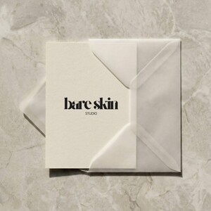 Peut inclure: Une carte cr&egrave;me avec le texte "bare skin STUDIO" en noir gras, plac&eacute;e dans une enveloppe translucide. L'enveloppe est carr&eacute;e et poss&egrave;de un rabat. Le fond est un marbre gris clair.
