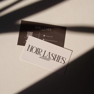 May include: Two business cards with the text "NOIR LASHES studio". One card is white with black text, the other is dark brown with white text and a QR code. The cards are on a white surface with shadows.