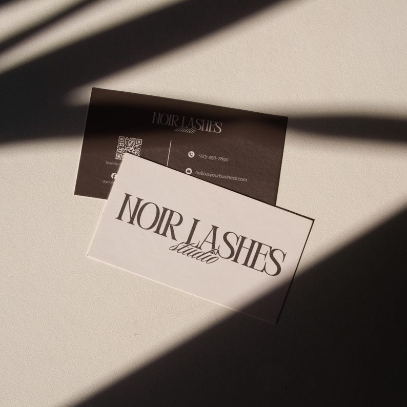 May include: Two business cards with the text "NOIR LASHES studio". One card is white with black text, the other is dark brown with white text and a QR code. The cards are on a white surface with shadows.