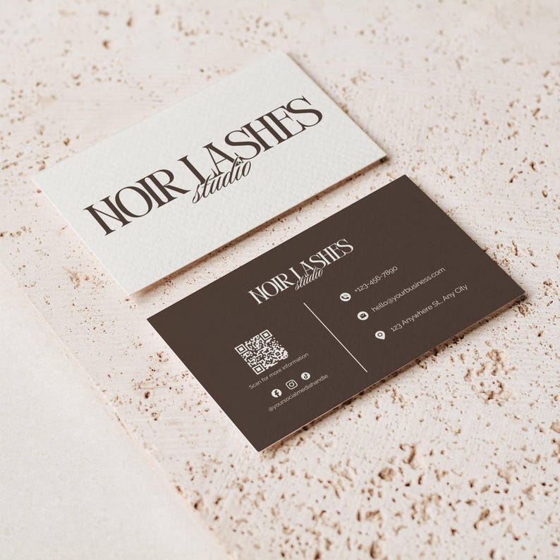 May include: Two business cards with the text "NOIR LASHES studio". One card is white with black text, the other is dark brown with white text and a QR code. The cards are on a textured, light beige surface.