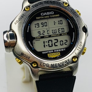 May include: Silver Casio Twin Sensor watch with a black digital display showing the time, date, and temperature. The watch has a black leather band and gold-colored accents. The watch is water resistant to 200m.