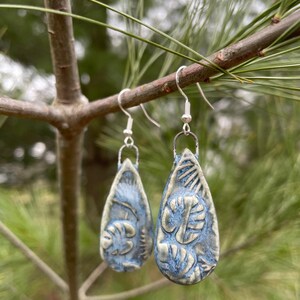 May include: A pair of handmade, blue ceramic earrings with a teardrop shape. Each earring features a raised, textured design of leaves and other organic shapes. The earrings hang from silver-colored hooks, displayed on a tree branch.