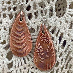 May include: A pair of handmade ceramic earrings. Each earring is a teardrop shape with a reddish-brown glaze and a botanical design. The earrings hang from silver-colored ear wires. The background is a cream-colored, open-weave fabric.