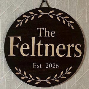 May include: A round, dark brown wooden sign with the text "The Feltners Est 2026" in a light beige color. The sign is decorated with a beige leaf design around the edge. A small metal loop is attached at the top for hanging.