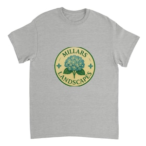 以下が含まれることがあります： ライトグレーのTシャツ。円形のロゴには「Millars Landscapes」の文字、青いアジサイの花、緑のアクセントが描かれています。柔らかい素材で作られたTシャツです。