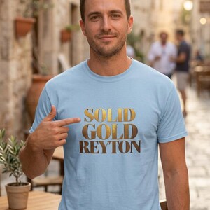 May include: Light blue t-shirt with the words "SOLID GOLD REYTON" in a gold metallic font. The shirt is being pointed at by a person. The background is a blurred outdoor scene with buildings and people.