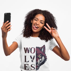 May include: White t-shirt with the word "WOLVES" in a maroon and white pattern, and a maroon and white wolf graphic. The person in the image is holding a black smartphone and making a peace sign.