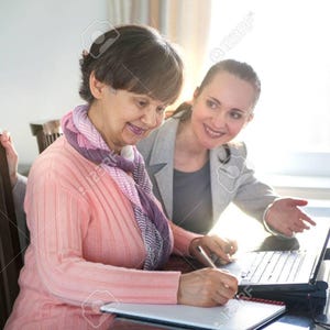 May include: Two women are seated at a table, one writing on a notepad while the other points at a laptop screen. The woman writing wears a pink sweater and a patterned scarf. The other woman wears a grey blazer. The scene is brightly lit.