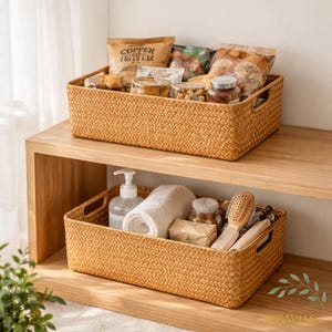 May include: Two woven rectangular storage baskets, one on top of the other, filled with various items. The top basket contains packaged food items and jars. The bottom basket holds toiletries, including a bottle of soap, a rolled towel, and a brush.