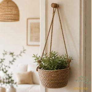 Puede incluir: Un macetero colgante de mimbre, suspendido por cuerdas de fibra natural de una clavija de madera. La cesta contiene plantas verdes. El fondo es de color neutro, con un enfoque suave en otra decoración.