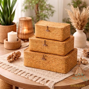 Puede incluir: Tres cajas de almacenamiento rectangulares tejidas de diferentes tamaños, apiladas sobre una mesa. Las cajas son de color beige claro con una textura tejida y cierres de madera. Forman parte de una colección de decoración del hogar.