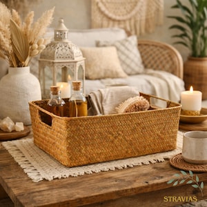 May include: A woven rectangular basket with handles, filled with spa essentials, sits on a wooden table. The basket contains small glass bottles, a washcloth, and a brush. A lit candle and decorative items complete the scene.