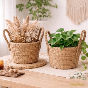 Może przedstawiać: Dwa plecione kosze z uchwytami z liny. Jeden kosz zawiera suszone kwiaty, drugi zielone rośliny. Oba stoją na drewnianym stole, z plecioną podkładką i elementami dekoracyjnymi.