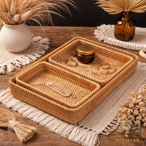 May include: Woven rattan tray set with a rectangular design. The set includes three trays of varying sizes, perfect for organizing small items. A small jar with a gold lid and jewelry are placed on the trays. The trays are on a beige macrame placemat.