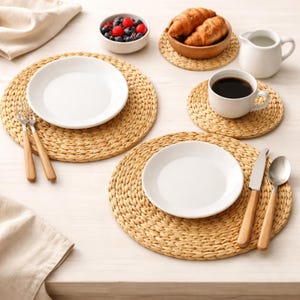 May include: A breakfast table setting featuring woven placemats, white plates, and cutlery with wooden handles. There's a bowl of berries, croissants, coffee, and a creamer. The round placemats are made of a light brown woven material.