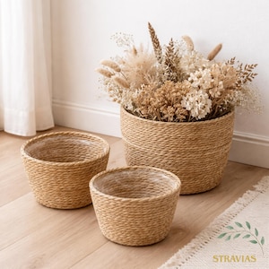 Könnte beinhalten: Drei geflochtene, hellbraune Blumentöpfe in verschiedenen Größen. Der größte Topf enthält einen Strauß Trockenblumen in Beige- und Brauntönen. Die Töpfe sind mit klarem Kunststoff ausgekleidet. Die Töpfe stehen auf einem hellen Holzboden.
