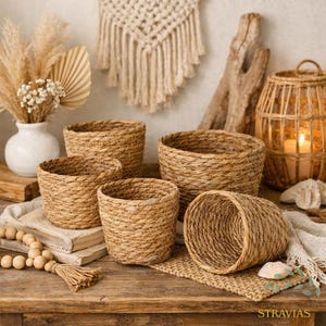 Puede incluir: Una colección de cestas cilíndricas tejidas de varios tamaños. Las cestas de tonos naturales están dispuestas sobre una superficie de madera, con un tapiz de macramé y un jarrón con flores secas en el fondo. Las cestas están hechas de un material texturizado de color marrón claro.