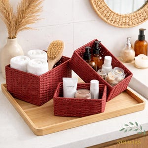 Peut inclure: Ensemble de trois paniers de rangement rouges tressés de différentes tailles, disposés sur un plateau en bois. Les paniers contiennent des serviettes blanches roulées, des articles de toilette et une brosse en bois. Un ensemble d'accessoires de salle de bain décoratifs.