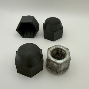 Puede incluir: Cuatro cubiertas negras para tuercas de rueda y una tuerca de rueda plateada. Tres de las cubiertas tienen forma de cúpula y una está abierta. La tuerca de rueda plateada tiene forma hexagonal con roscas visibles.
