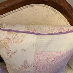 May include: A decorative cushion with a patchwork design featuring floral and patterned fabrics in cream, pink, and lavender. The cushion has a lavender zip closure and is placed on a floral patterned chair.