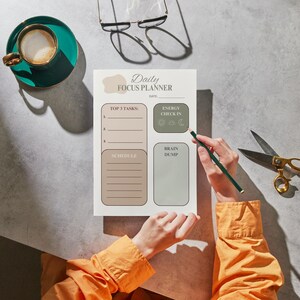 May include: A person writing on a "Daily Focus Planner" with a pencil. The planner has sections for tasks, schedule, and energy check-in. A teal cup of coffee, gold scissors, and glasses are also on the table.
