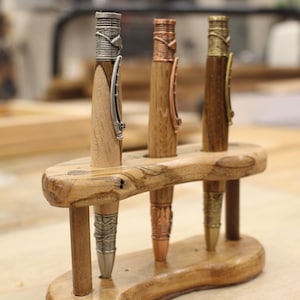 May include: Three handcrafted wooden pens with metal accents are displayed in a wooden pen holder. The pens have different metal finishes: silver, copper, and gold. The pen holder is made of light-colored wood.