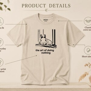 Puede incluir: Una camiseta beige de manga corta y cuello redondo con un estampado gr&aacute;fico negro de un conejo descansando. El texto debajo del conejo dice, the art of doing nothing. La camiseta presenta un corte unisex y detalles del producto visibles.