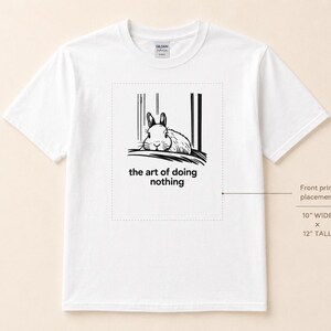 Puede incluir: Una camiseta blanca de manga corta con un gr&aacute;fico negro de un conejo descansando y el texto "the art of doing nothing". Un diagrama indica que el &aacute;rea de impresi&oacute;n frontal mide 25,4 cm de ancho por 30,48 cm de alto.