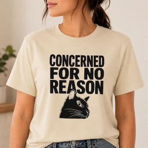 May include: A cream-colored short-sleeve t-shirt featuring the black text CONCERNED FOR NO REASON printed in a bold sans-serif font. Below the text is a black and white graphic illustration of a cat head with wide, alert eyes.