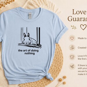 Puede incluir: Una camiseta de manga corta de color azul claro con un gr&aacute;fico negro de un conejo descansando, acompa&ntilde;ado por el texto "the art of doing nothing" impreso debajo de la ilustraci&oacute;n. La prenda tiene cuello redondo y un corte est&aacute;ndar.