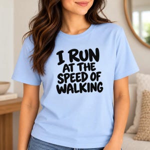 May include: A light blue short-sleeve t-shirt featuring the black text "I RUN AT THE SPEED OF WALKING" printed in a bold, rounded font across the chest. The garment has a classic crew neckline and a straight hemline, providing a simple design.