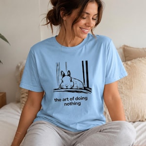 Puede incluir: Una camiseta de manga corta de color azul claro con una ilustraci&oacute;n gr&aacute;fica negra de un conejo descansando entre dos l&iacute;neas verticales. Debajo del conejo, el texto dice "the art of doing nothing" en una tipograf&iacute;a negra sencilla.