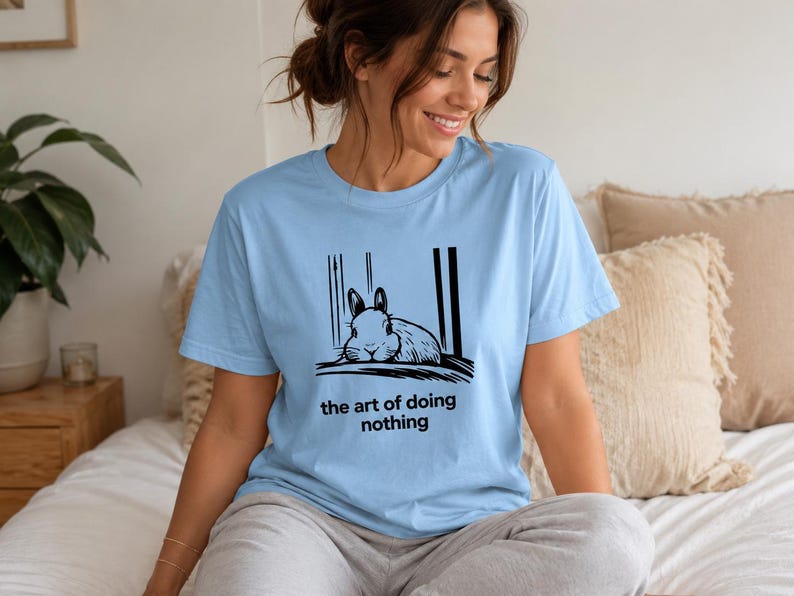 Puede incluir: Una camiseta de manga corta de color azul claro con una ilustraci&oacute;n gr&aacute;fica negra de un conejo descansando entre dos l&iacute;neas verticales. Debajo del conejo, el texto dice "the art of doing nothing" en una tipograf&iacute;a negra sencilla.