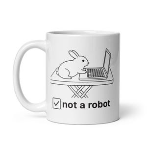 Könnte beinhalten: Weiße Keramiktasse mit einer Schwarz-Weiß-Illustration eines Kaninchens, das einen Laptop benutzt. Der Text "not a robot" ist unter dem Bild mit einem Häkchen in einem Kästchen aufgedruckt.