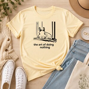 Puede incluir: Una camiseta de manga corta de color amarillo p&aacute;lido con una ilustraci&oacute;n de l&iacute;neas negras de un conejo descansando y el texto "the art of doing nothing" impreso en una fuente sans-serif sencilla en el centro de la prenda.