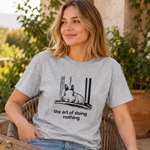 Puede incluir: Una camiseta gris jaspeado de manga corta con un estampado gr&aacute;fico negro de un conejo descansando sobre una superficie con l&iacute;neas verticales de fondo. Debajo de la ilustraci&oacute;n, el texto dice the art of doing nothing en letra negra.