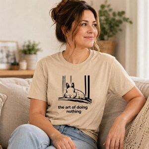 Puede incluir: Una camiseta beige de manga corta con un gr&aacute;fico negro de un conejo descansando y el texto "the art of doing nothing" impreso en la parte delantera. La prenda cuenta con un cuello redondo y un corte est&aacute;ndar de dise&ntilde;o sencillo.
