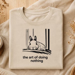 Puede incluir: Una camiseta beige de cuello redondo con una ilustraci&oacute;n de l&iacute;neas negras de un conejo descansando entre dos l&iacute;neas verticales. Debajo del gr&aacute;fico, el texto dice "the art of doing nothing" en una fuente negra sencilla y min&uacute;scula.