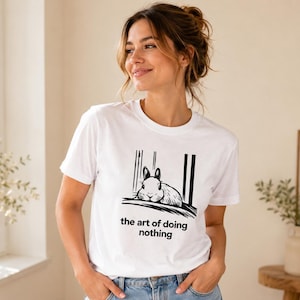 Puede incluir: Una camiseta blanca de manga corta con una ilustraci&oacute;n gr&aacute;fica negra de un conejo descansando sobre una superficie con l&iacute;neas verticales de fondo. Debajo de la ilustraci&oacute;n, el texto dice "the art of doing nothing" en letra negra.
