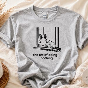 Puede incluir: Una camiseta gris jaspeado con una ilustraci&oacute;n gr&aacute;fica en negro de un conejo descansando. Debajo del conejo, el texto dice, the art of doing nothing. La camiseta tiene cuello redondo y mangas cortas, con un dise&ntilde;o sencillo y casual.