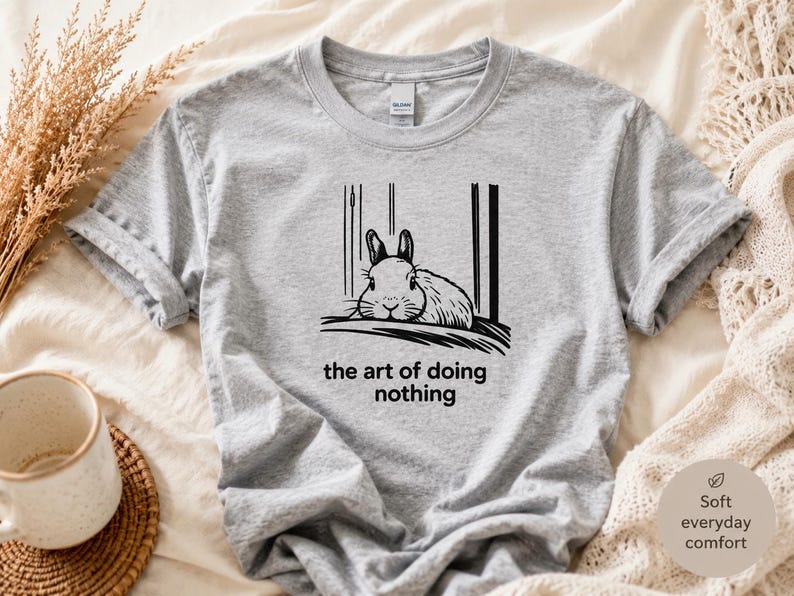Puede incluir: Una camiseta gris jaspeado con una ilustraci&oacute;n gr&aacute;fica en negro de un conejo descansando. Debajo del conejo, el texto dice, the art of doing nothing. La camiseta tiene cuello redondo y mangas cortas, con un dise&ntilde;o sencillo y casual.